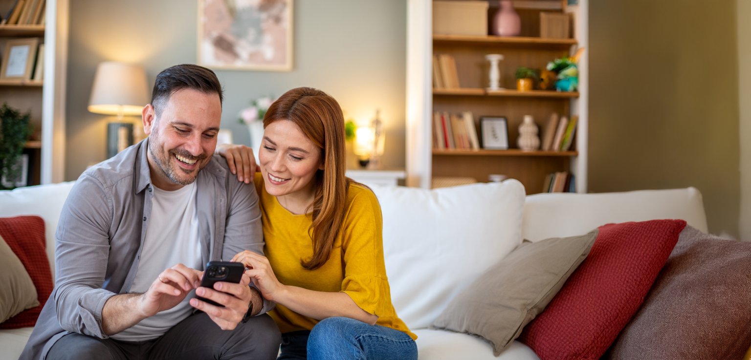 Ein lachendes Paar schaut gemeinsam auf ein Smartphone im gemütlichen Wohnzimmer.