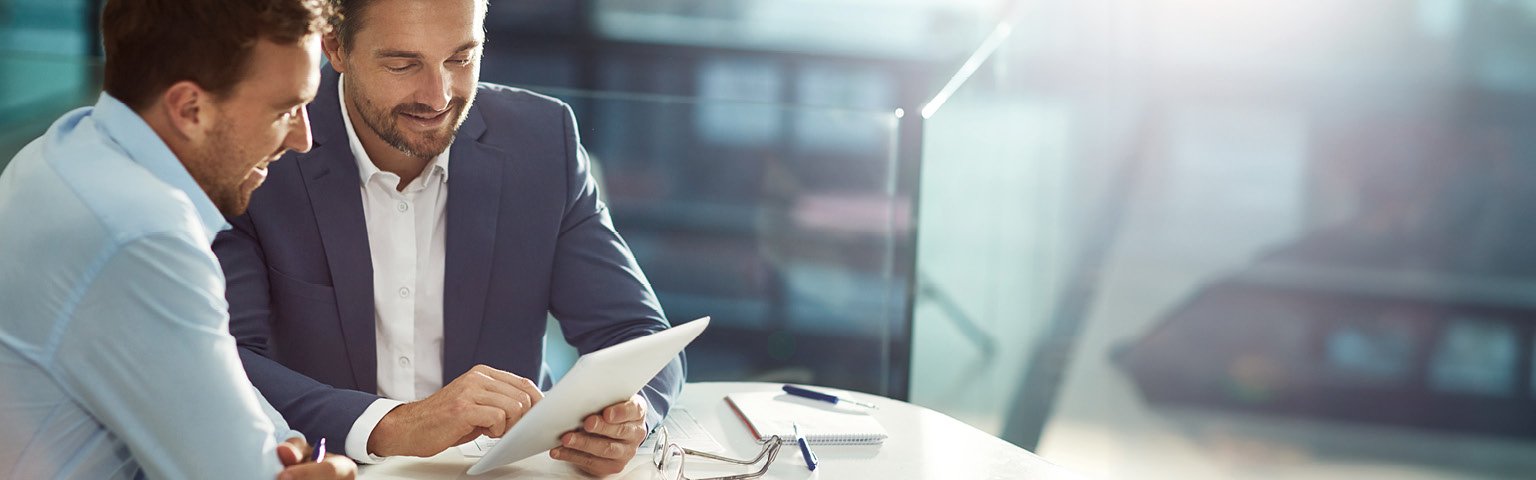 Zwei Männer in Anzug diskutieren über Dokumente an einem Tisch mit modernem Ambiente.