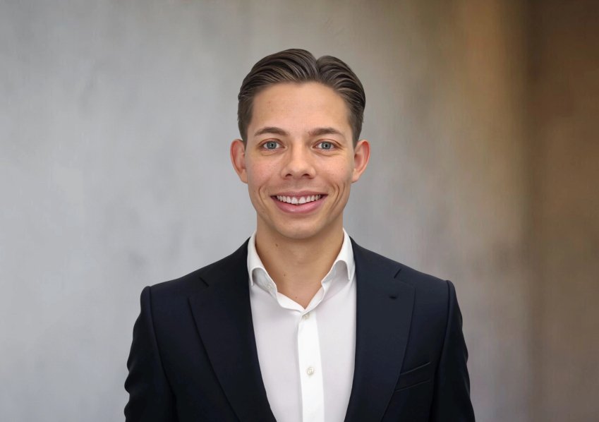 Ein lächelnder Mann in dunklem Anzug vor neutralem Hintergrund, professionell und freundlich.