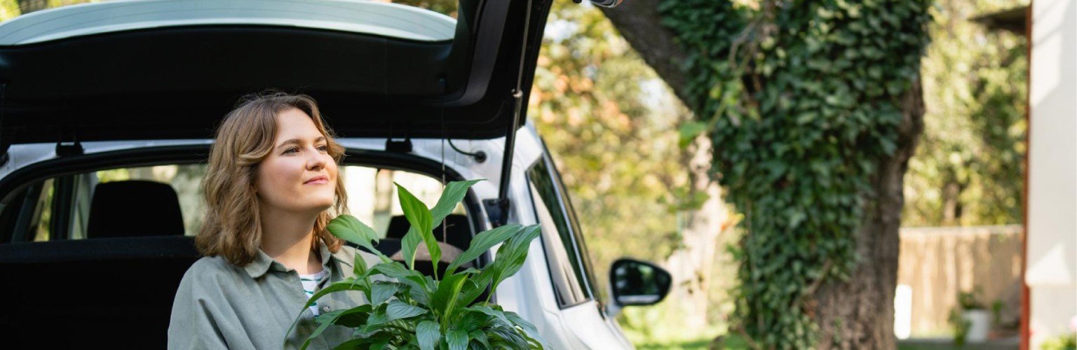 Frau mit Pflanze im Topf neben einem ladenden Elektroauto im Hof ​​eines Landhauses.