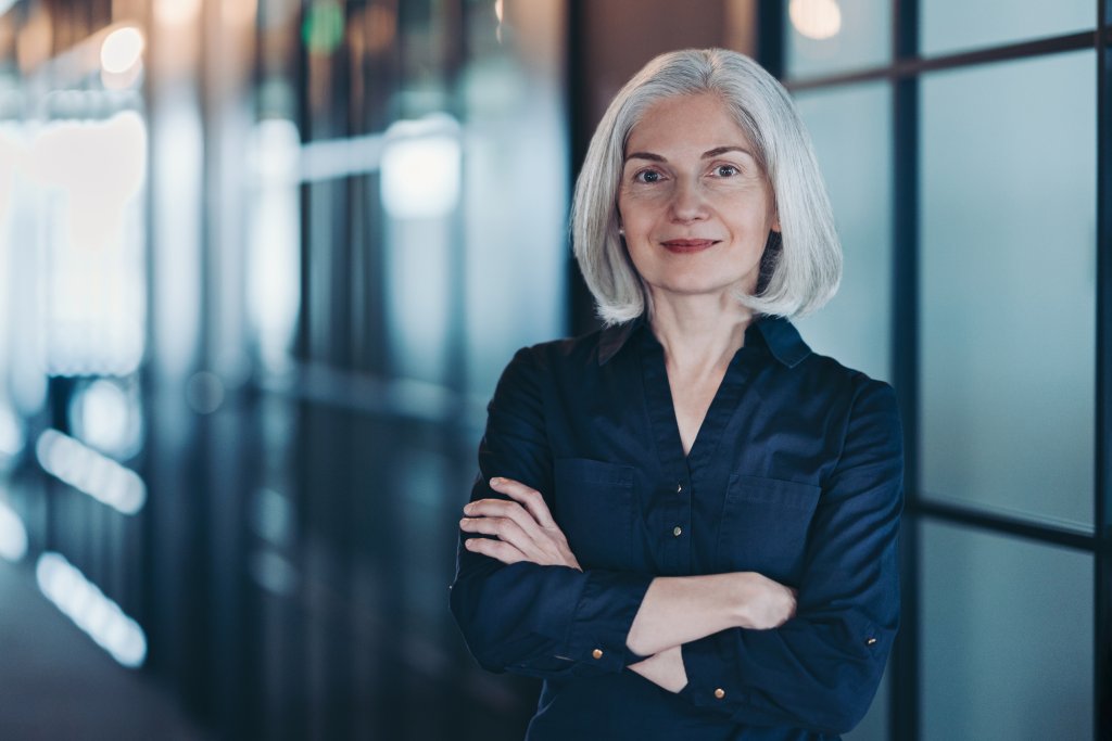 Selbstbewusste Frau mit grauem Haar steht mit verschlossenen Armen in einem modernen Bürogang.