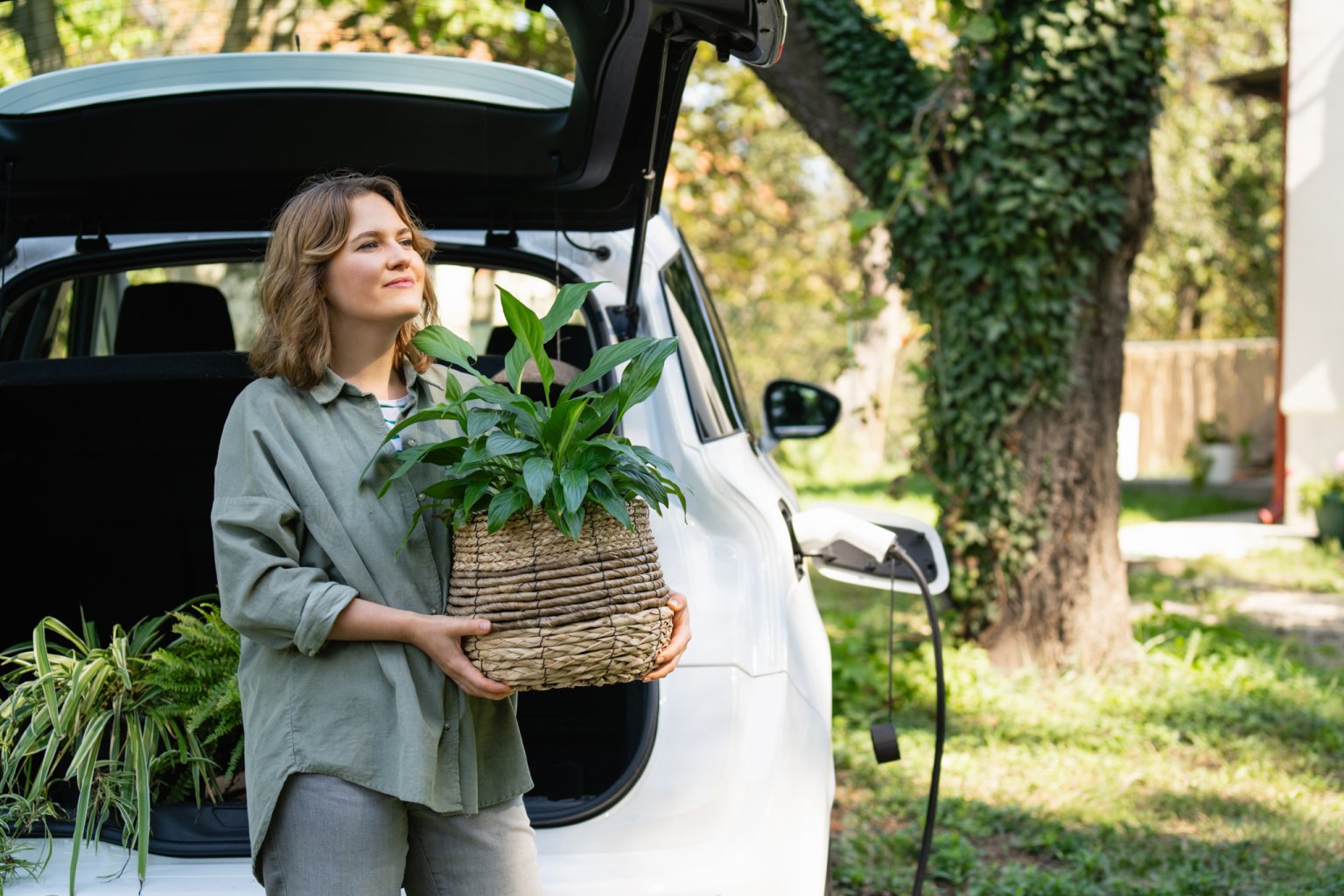 Eine Frau mit einer Pflanze steht am Kofferraum eines Autos. Das E-Auto wird aufgeladen aus dem Artikel Gut vorbereitet ins neue Zuhause – unsere Zügeltipps