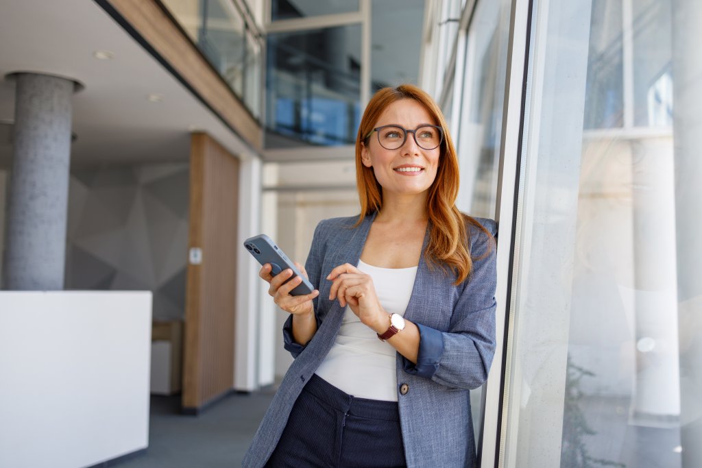 Frau mit Brille, in Businesskleidung, hält Smartphone und steht vor Fenster im Gebäude