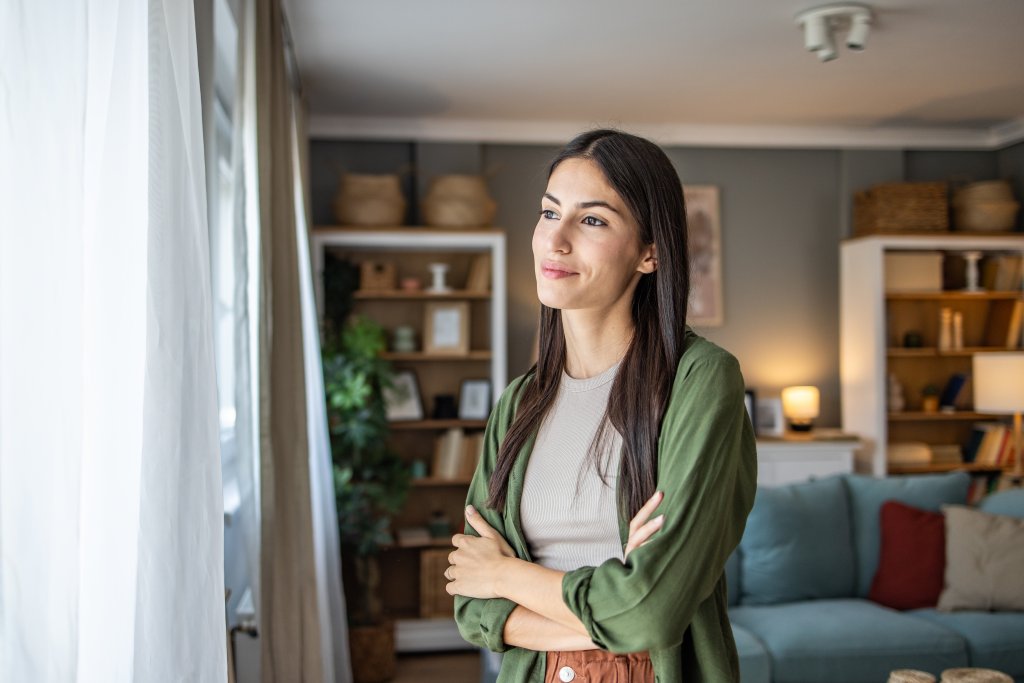 Frau steht vor einem Fenster im Wohnzimmer mit Bücherregalen im Hintergrund.