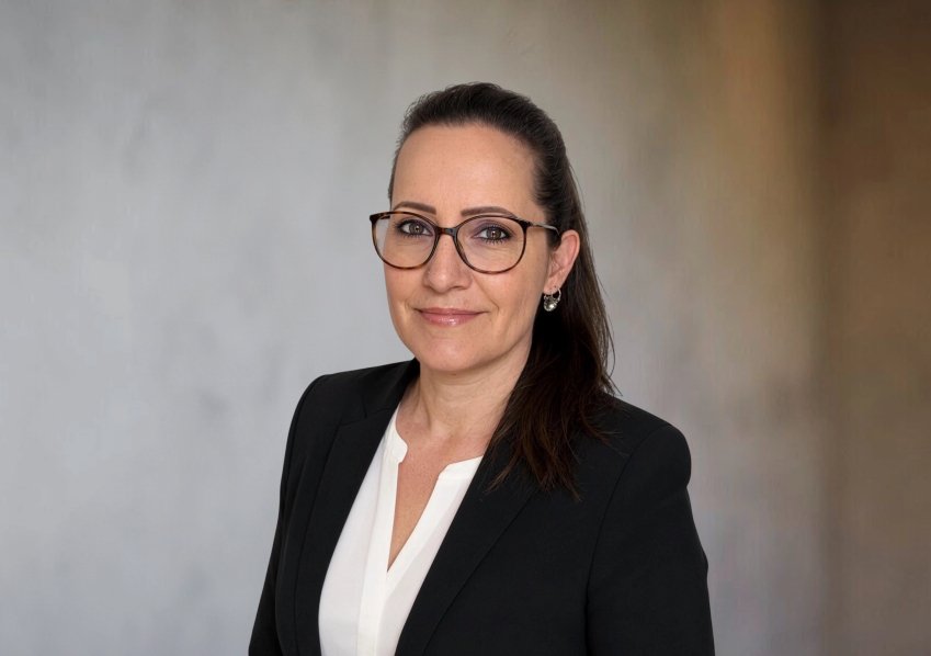 Frau in Businesskleidung mit Brille vor neutralem Hintergrund, professionell wirkend