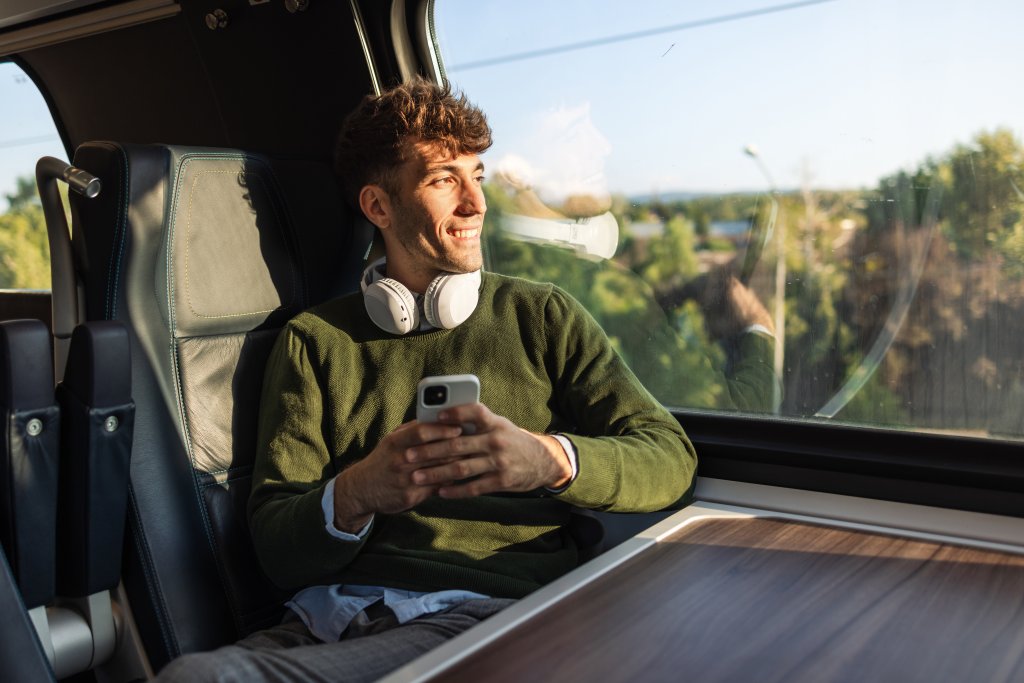 Ein junger Mann sitzt an einem Tisch im Zug, hält ein Smartphone und trägt Kopfhörer.