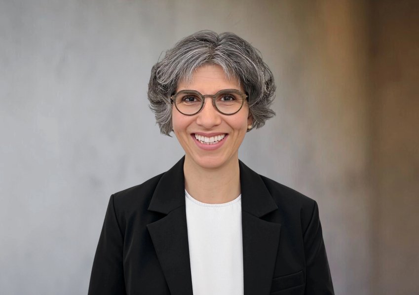 A smiling woman with gray hair, glasses, dark blazer and white shirt in front of neutral background