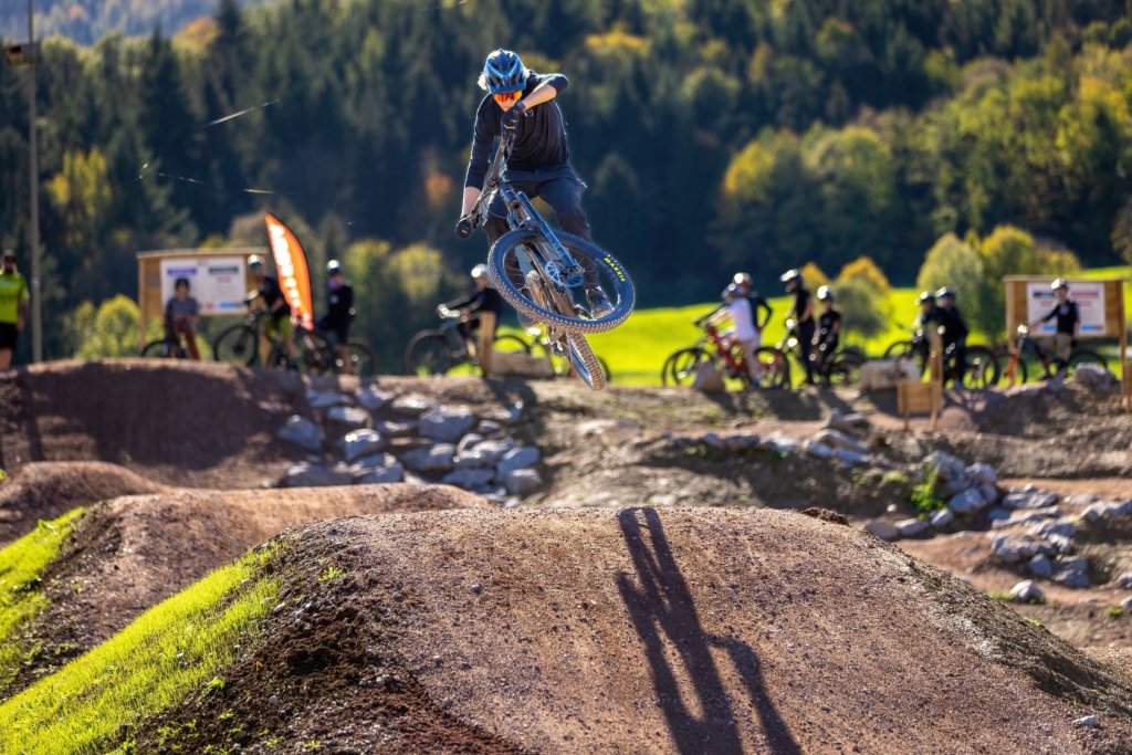 Mountainbiker in der Luft auf einem anspruchsvollen Trail im Grünen, im Hintergrund Biker und Natur.