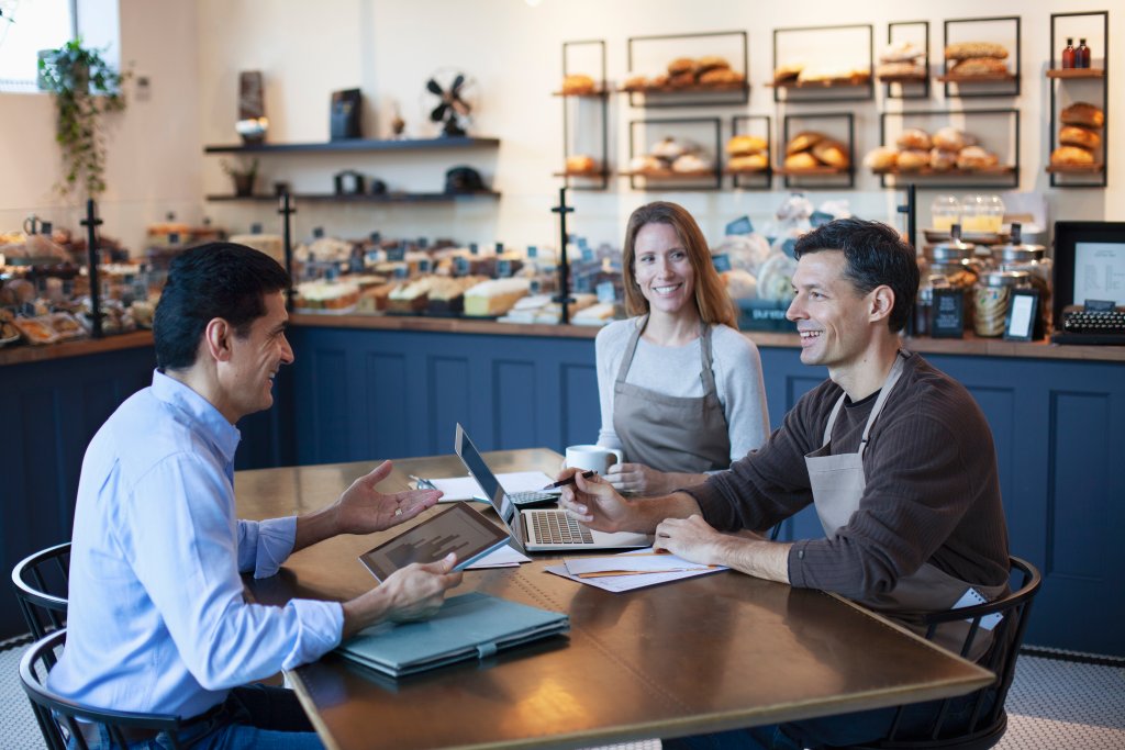Ein Gruppentreffen in einer Bäckerei mit zwei Personen, die plaudern, und einem Barista, der Kaffee serviert.