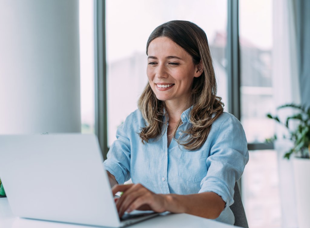 Frau arbeitet an einem Laptop in hellem Büro mit Fensterblick und Pflanzen.