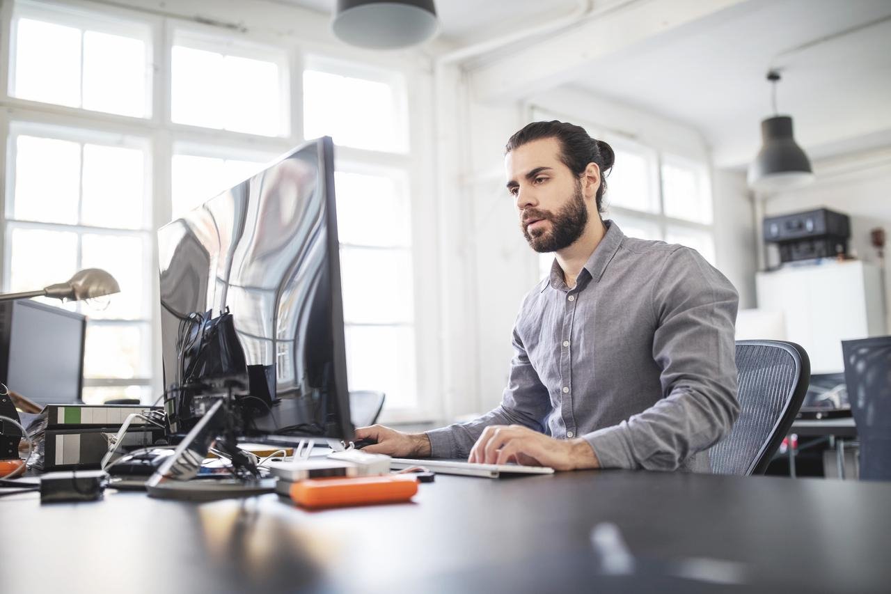 Mann arbeitet konzentriert am Computer in modernem Büro mit vielen Fenstern.