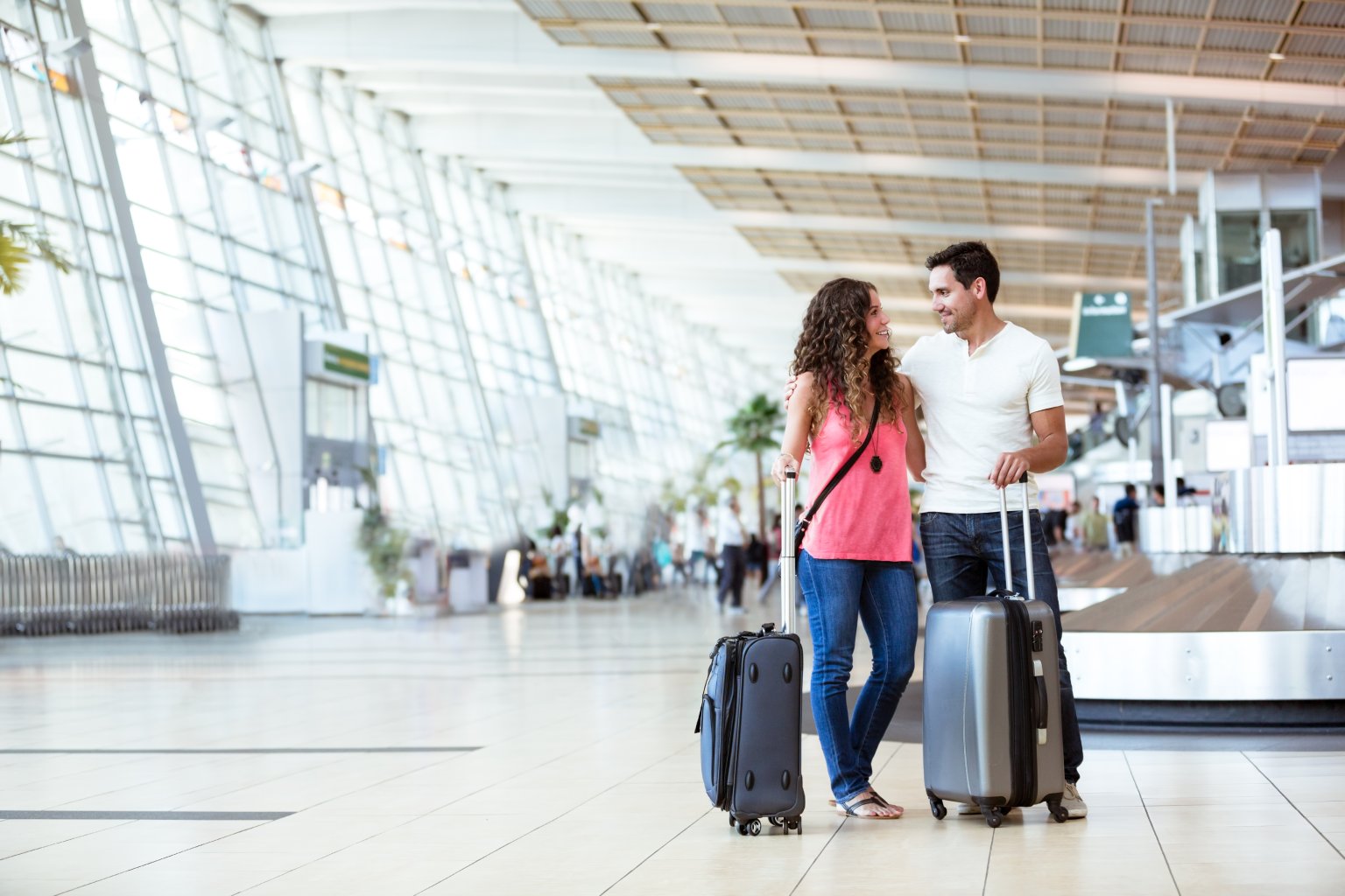 Zwei Personen mit Rollenkoffern stehen im Flughafen, im Hintergrund Glaswände und Check-in-Schalter. aus dem Artikel Beim Einkaufen oder Reisen: So profitieren Sie mit Ihrer Kreditkarte