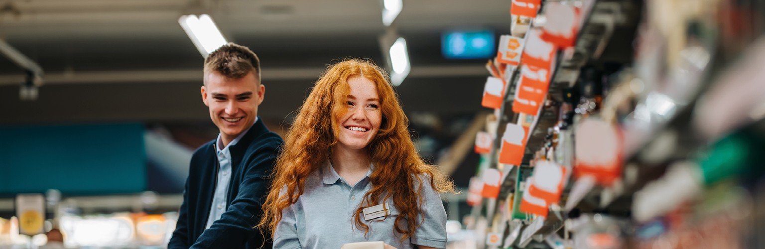 Zwei junge Mitarbeiter stellen Regale in einem Lebensmittelladen neu auf, lächelnd und engagiert in ihrer Arbeit.