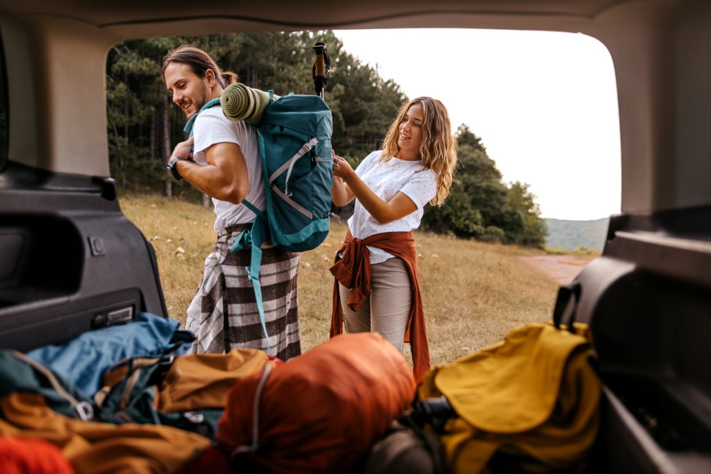 Zwei Menschen beim Camping mit Rucksack, im offenen Kofferraum, Naturhintergrund.