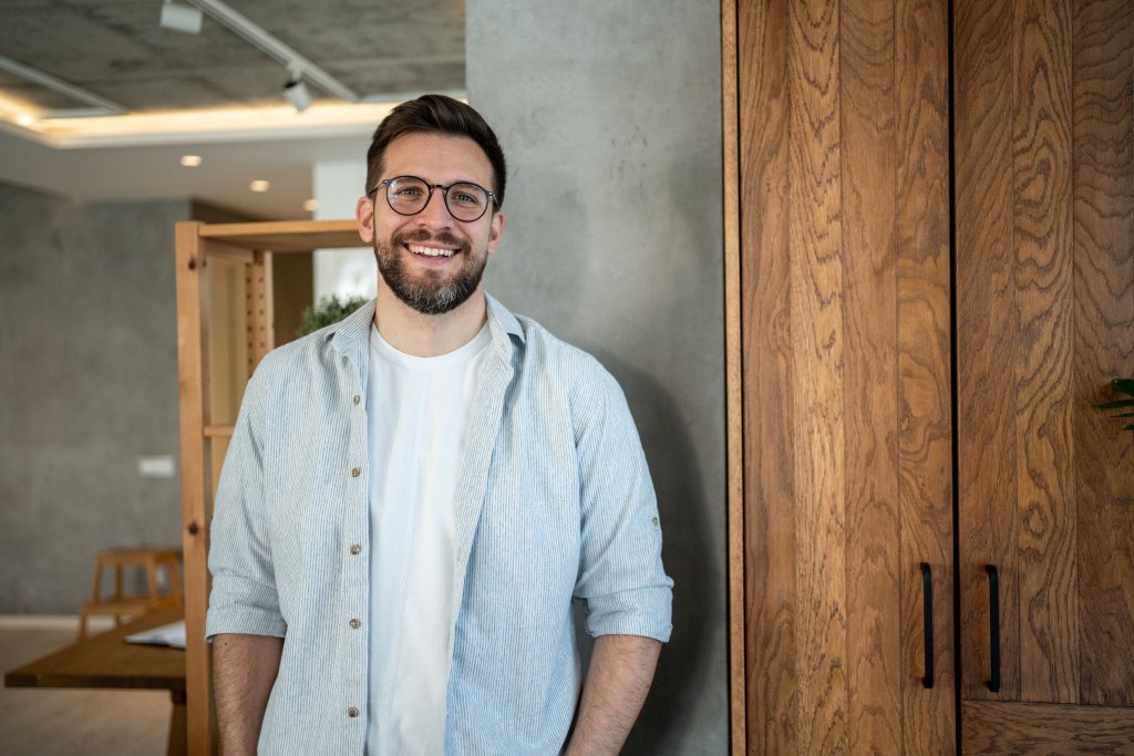 Mann mit Brille, helles Hemd, lächelt, steht vor Holzschrank im Büro.