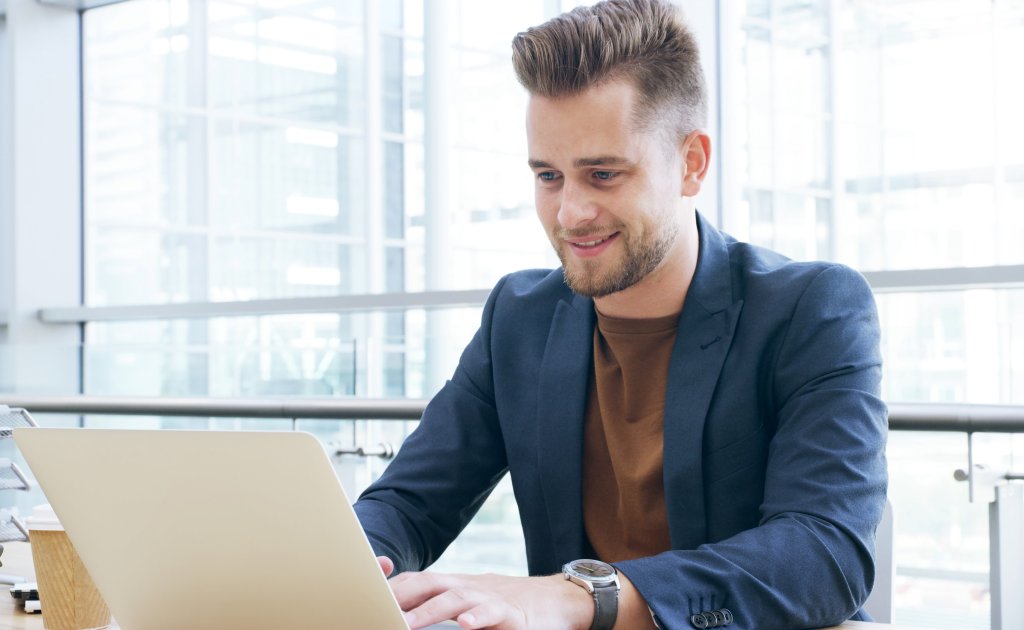 Ein Mann sitzt mit einem Laptop in einem hellen Büro mit großen Fenstern.