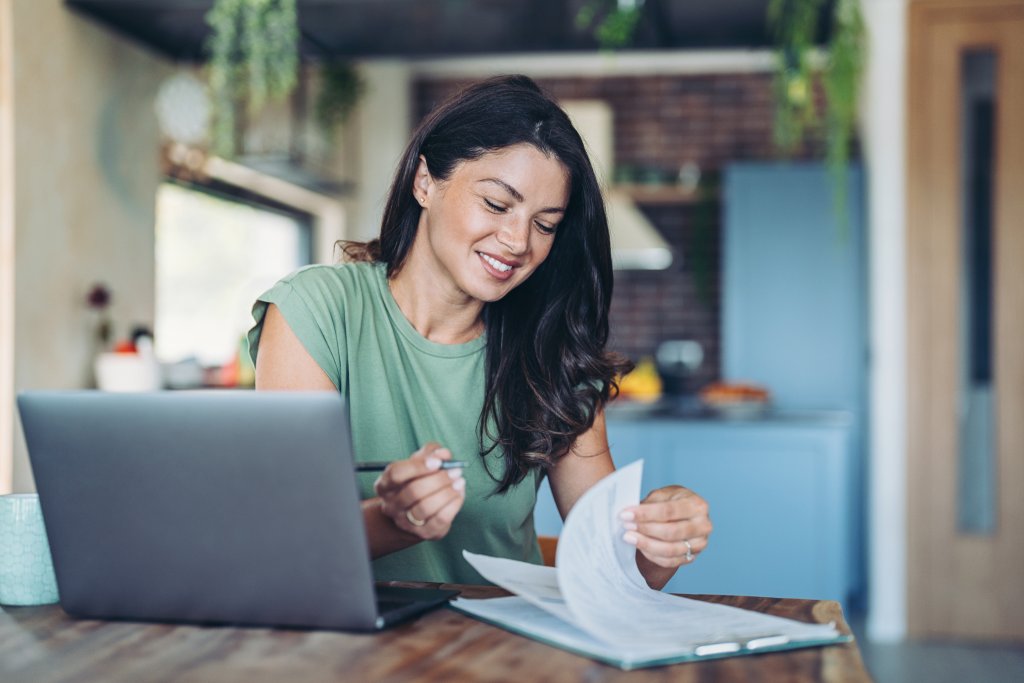 Frau arbeitet an Dokumenten mit einem Laptop, lächelnd in einem gemütlichen Homeoffice-Setting.