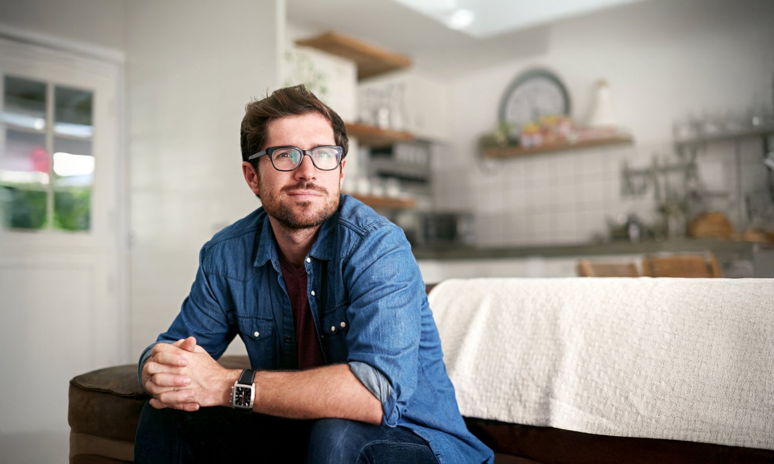 Ein Mann mit Brille und Jeans sitzt auf einer Couch in einer modernen Wohnung.