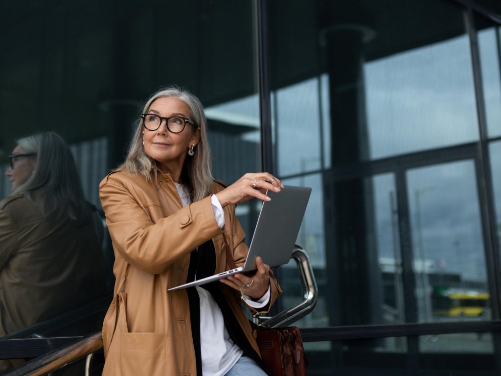 Ältere Frau steht mit einem Laptop vor einem modernen Gebäude.