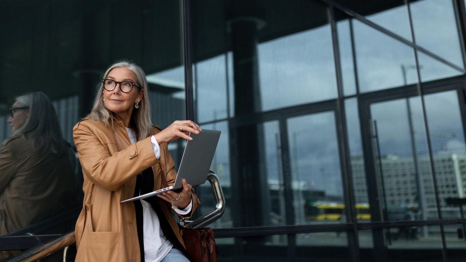Ältere Frau steht mit einem Laptop vor einem modernen Gebäude.