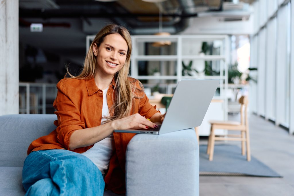 Eine Frau sitzt auf einem Sofa vor einem Laptop in einem modernen Büro mit viel Tageslicht.