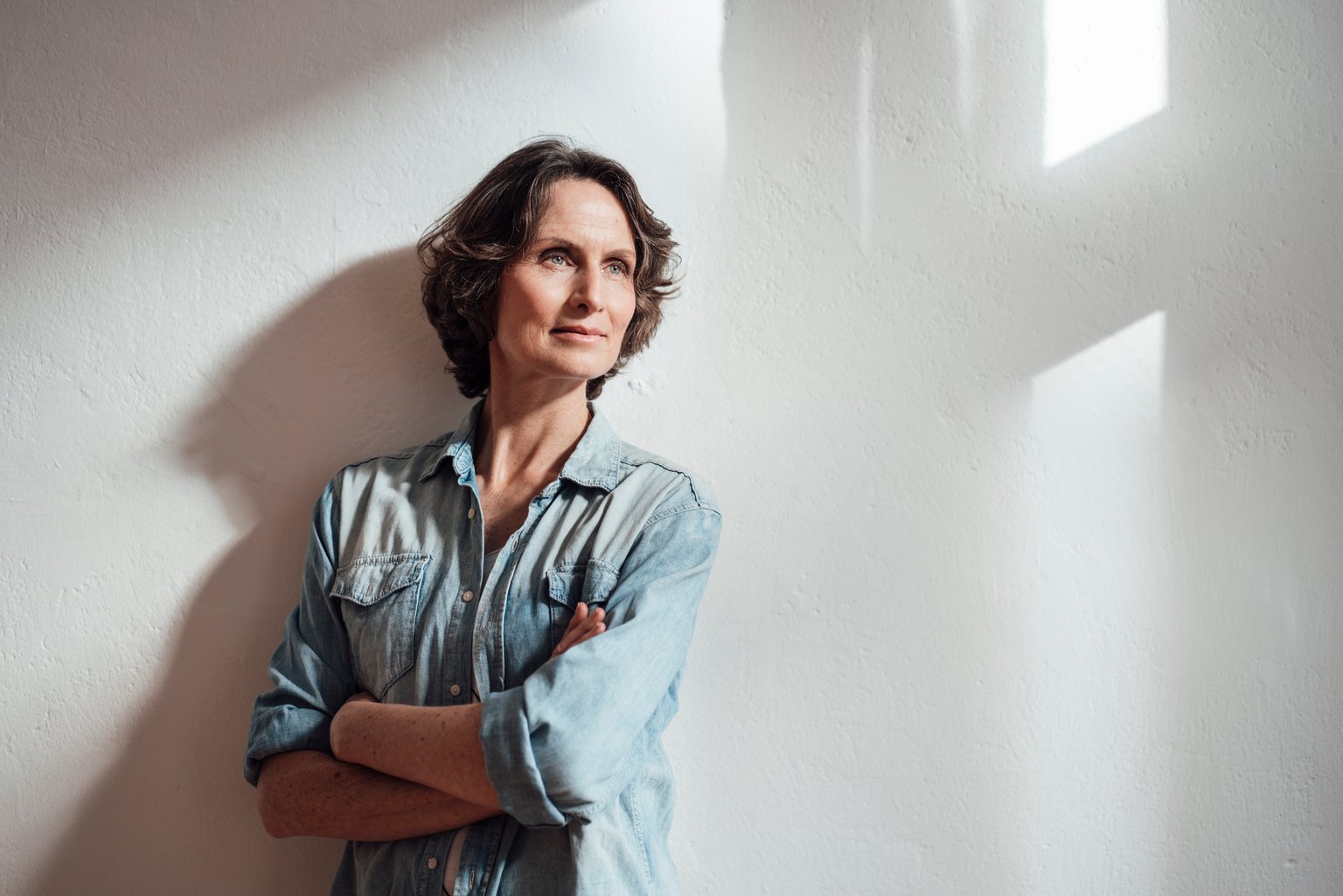 Eine Frau mit kurzen Haaren, die ein Denim-Hemd trägt, steht an einer sonnenbeschienenen Wand und schaut nachdenklich voraus. aus dem Artikel Nachhaltig anlegen – die drei grössten Mythen