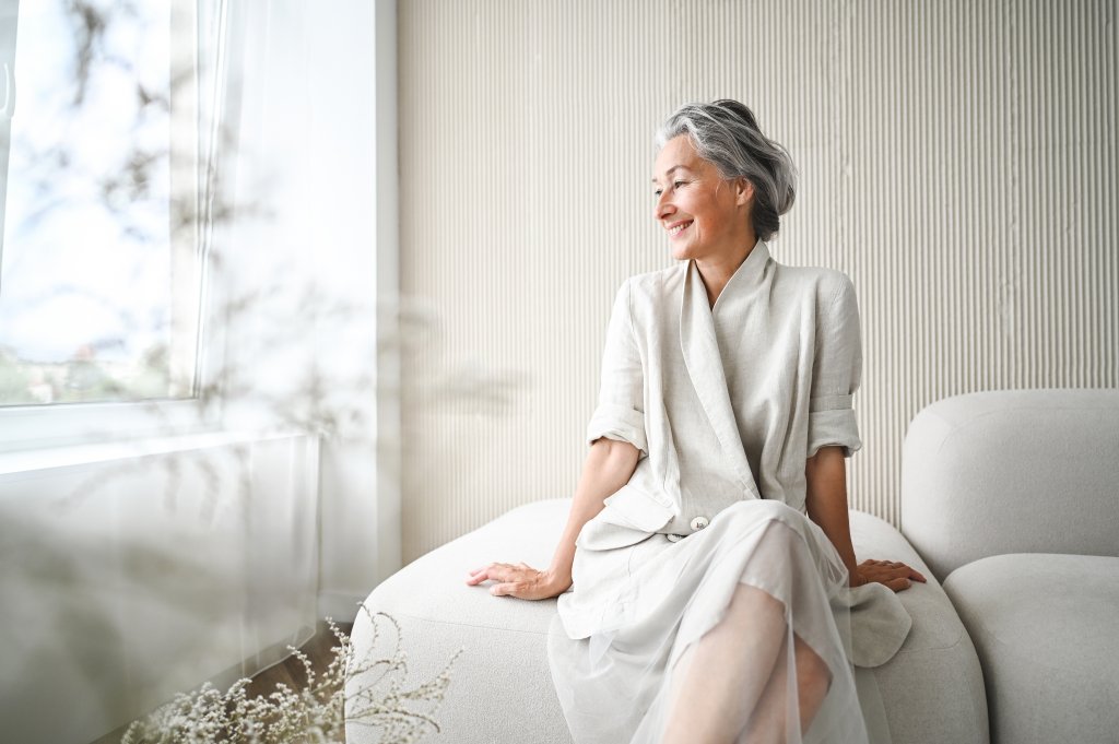 Frau sitzt auf einem Sofa in hellen Kleidung vor einem hellen Hintergrund.