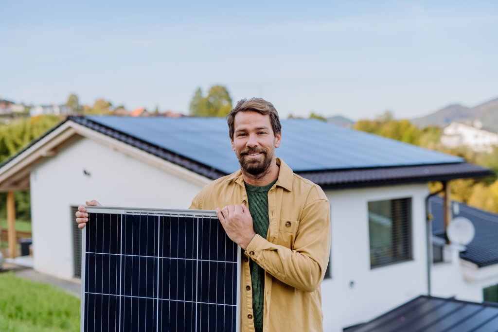 Ein älterer Mann hält ein Solarpanel in der Nähe seines Hauses, auf dessen Dach sich Solarpaneele befinden. Konzept für alternative Energien, Ressourcenschonung und nachhaltigen Lebensstil.