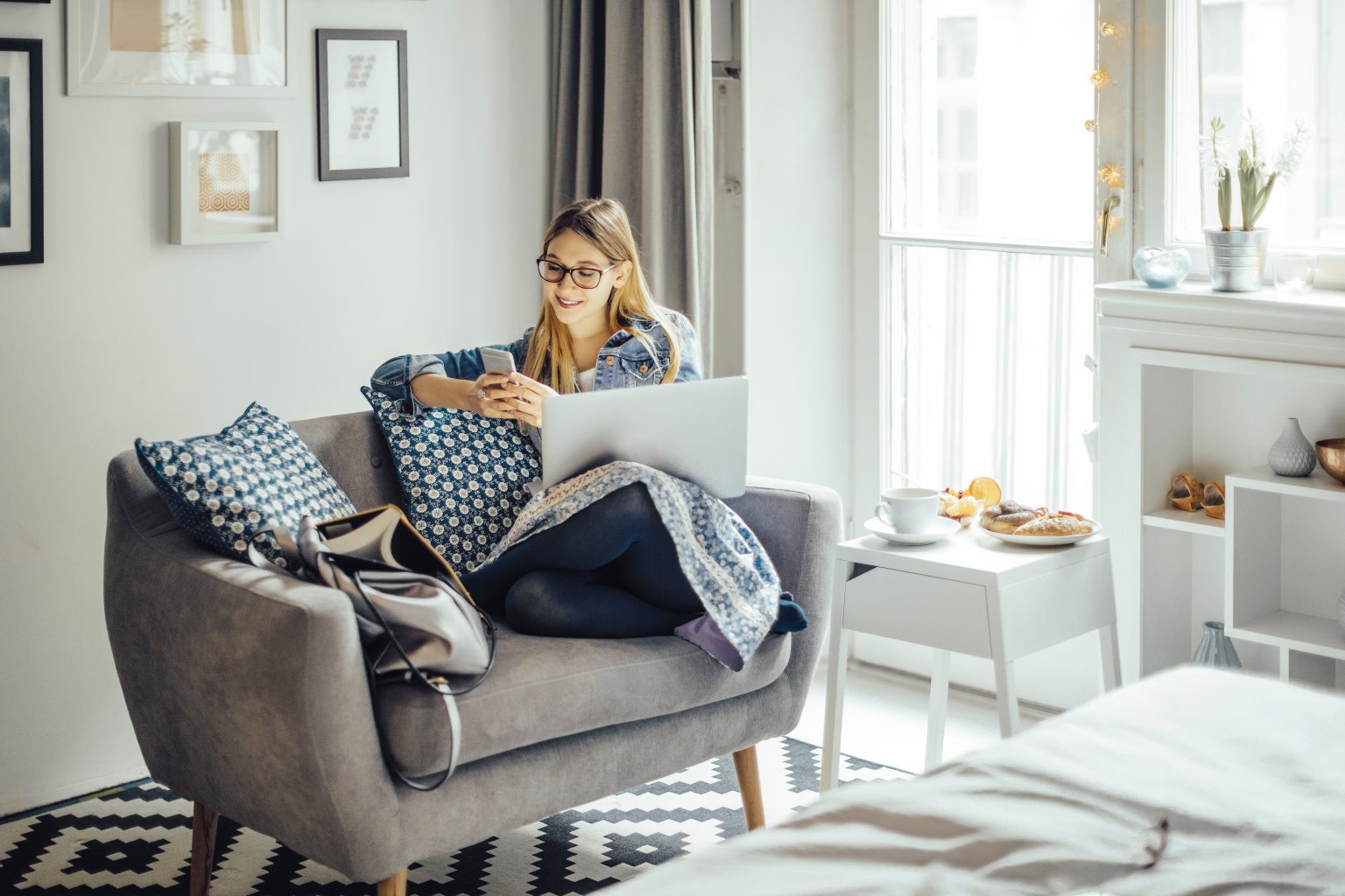 Frau mit Laptop und Handy in modernem Wohnzimmer mit gemütlicher Atmosphäre.