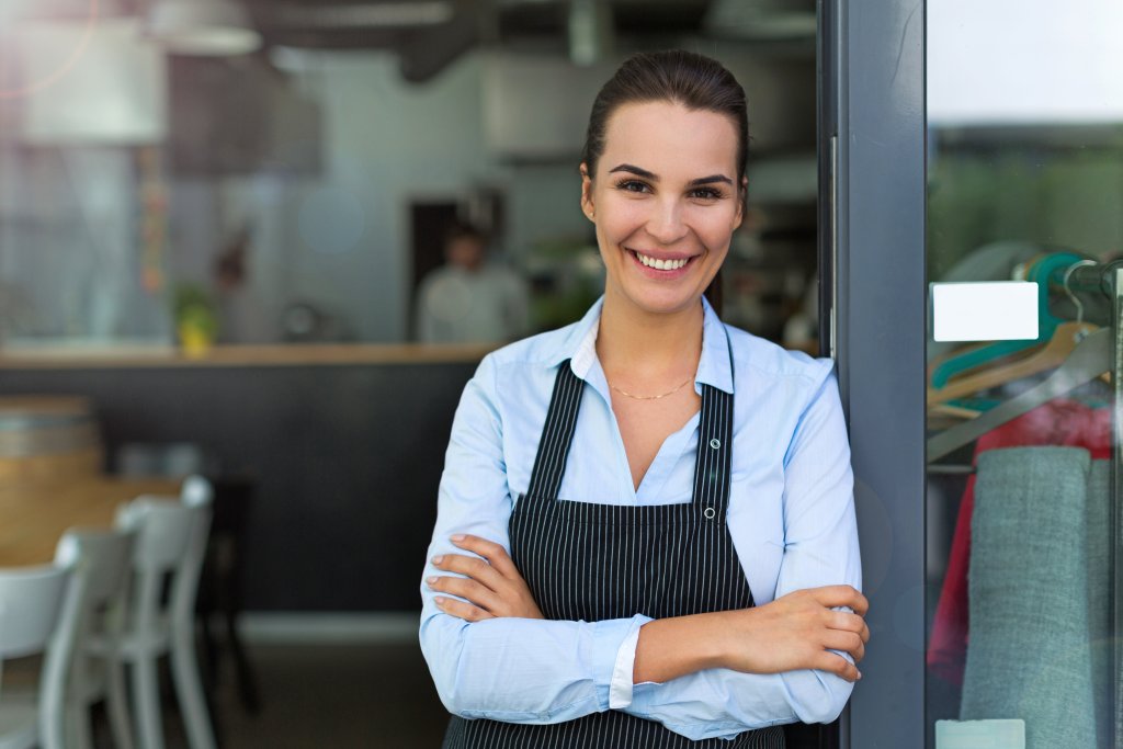 Eine lächelnde Frau in einer Schürze steht selbstbewusst am Eingang eines Cafés.