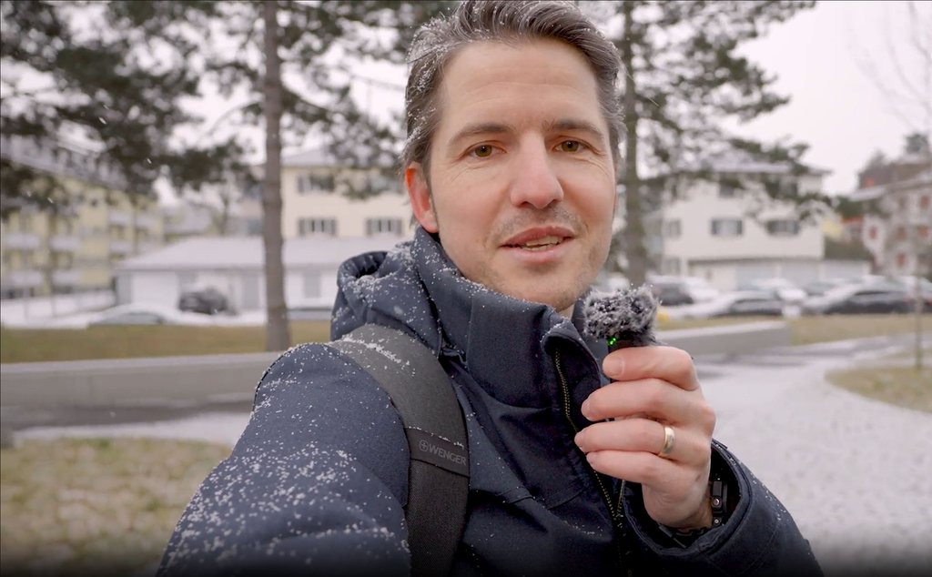 Person hält Smartphone vor das Gesicht, trägt dunkle Jacke und Rucksack.