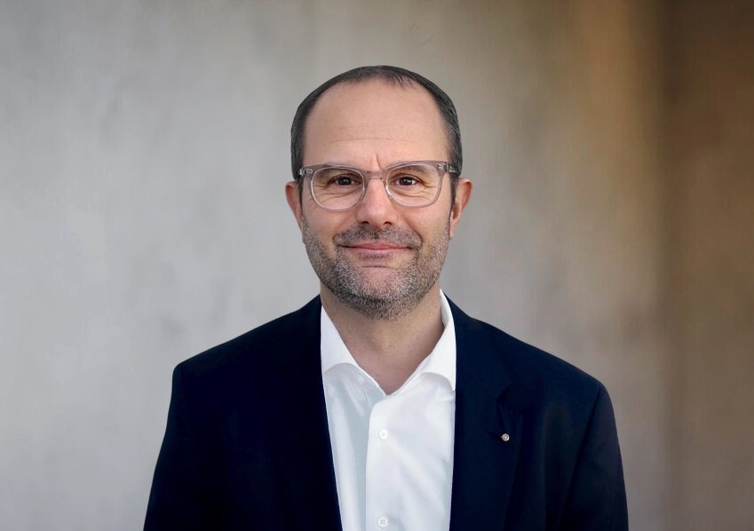 Ein Mann mit Brille und Bart trägt dunklen Anzug und steht vor neutralem Hintergrund.
