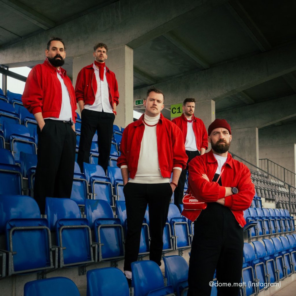 Fünf junge Menschen in roten Jacken stehen auf blauen Stadionbänken im Untergeschoss.