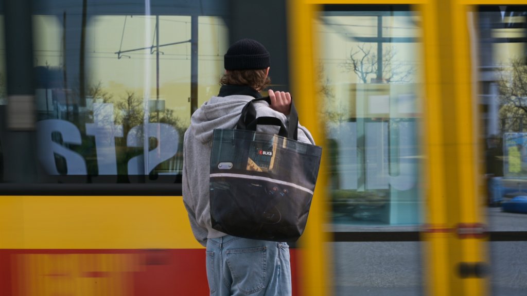 Ein junger Mann steht mit einer Unikat-Tasche an einer Bushaltestelle vor einem gelben Bus.