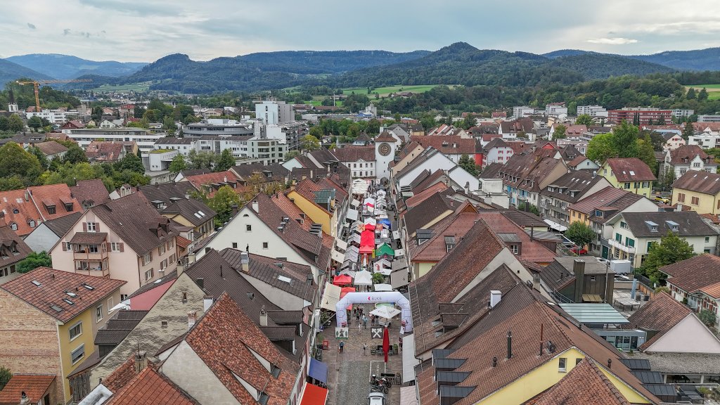 Lange Tafel in Laufen. Strassen mit Zelten und Menschen, umgeben von Gebäuden und Hügellandschaft.