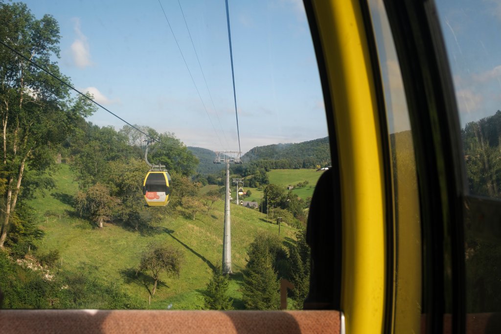 Seilbahn fährt über grüne Hügel mit Bäumen und weiten Feldern in blauer Himmelswelt