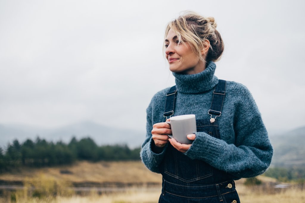 Frau mit Tasse in der Natur, umgeben von sanften Hügeln und grauem Himmel.