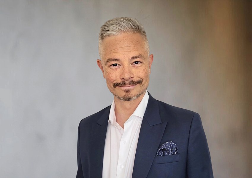 Ein Mann in dunklem Anzug mit weißem Hemd, Hintergrund grau und beige.
