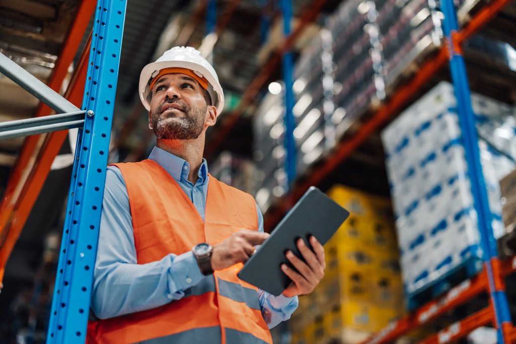Mann in Orange Weste mit Tablet in einem Lager mit hohen Regalen