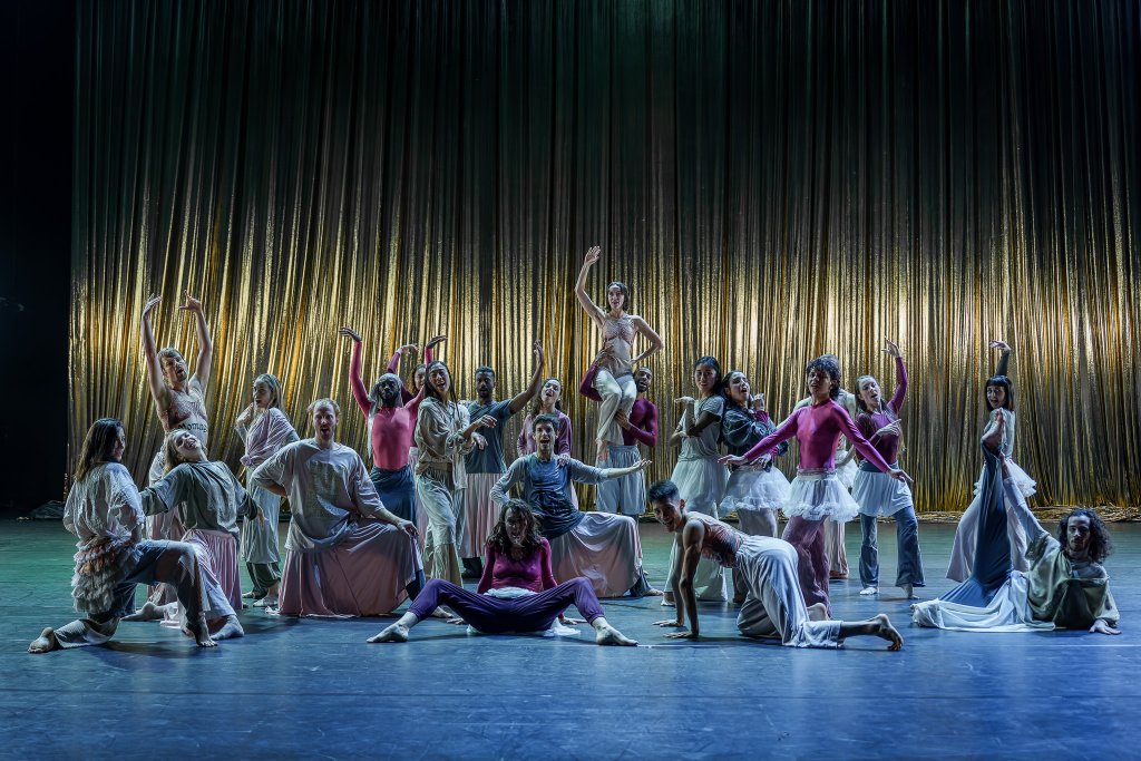 Tanzende Menschen in bunten Kostümen vor einem goldenen Vorhang auf der Bühne.