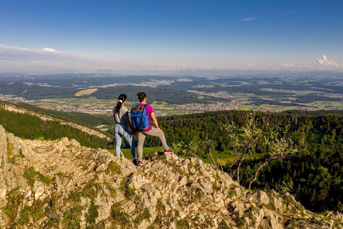 Zwei Wanderer auf einem Berggipfel mit Blick auf die grüne Landschaft und den blauen Himmel.