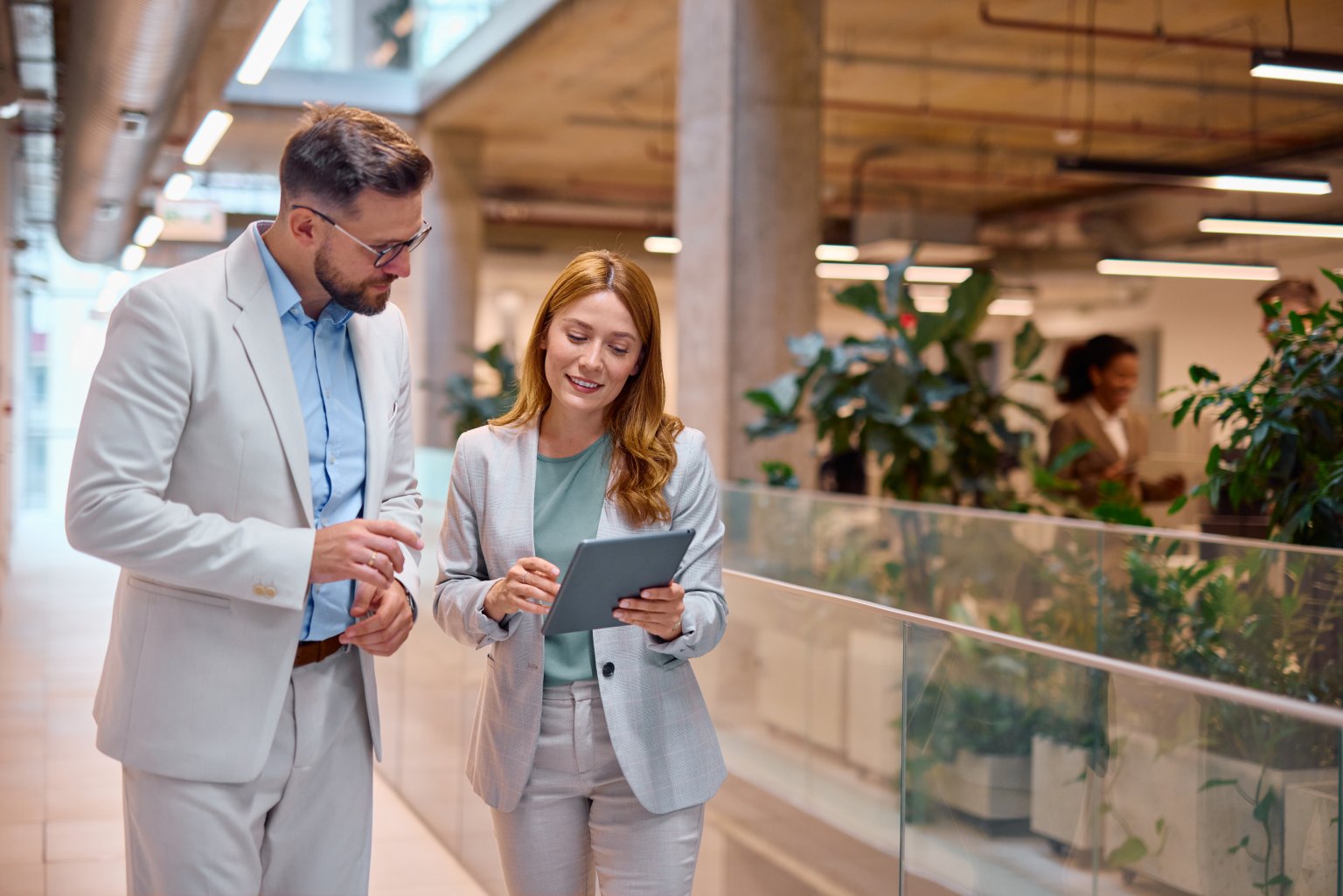 Zwei Geschäftsleute gehen durch ein modernes Büro, besprechen ihre Arbeit und arbeiten in einer hellen, umweltfreundlichen Umgebung mit einem Tablet an einem Projekt zusammen.