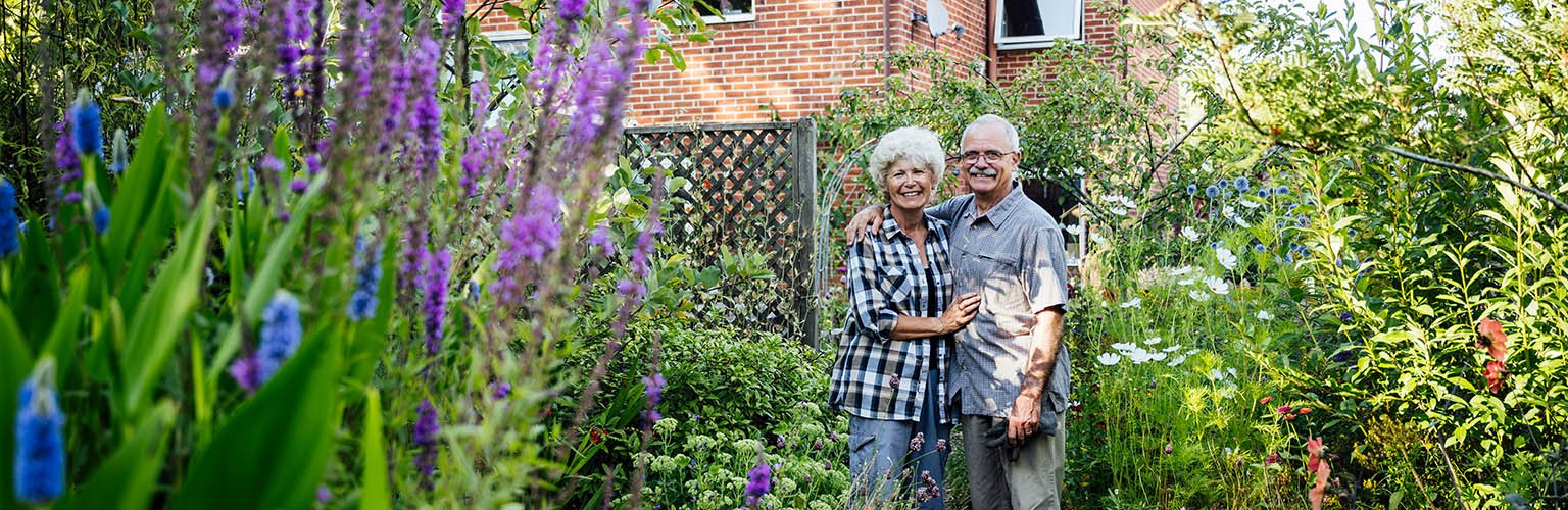 Ein älteres Paar lächelt glücklich in einem blühenden Garten vor ihrem Haus.
