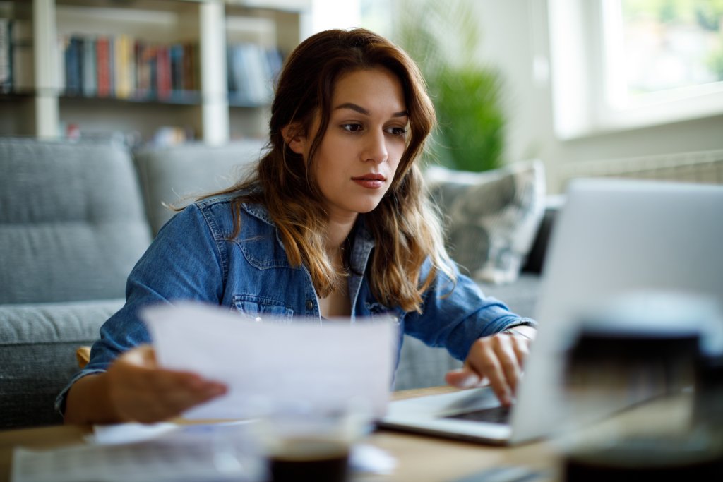 Frau arbeitet konzentriert an ihrem Laptop und prüft Unterlagen in einem modernen Wohnraum.