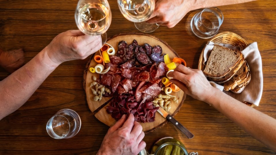 Mehrere Hände halten Weingläser über einer Käse- und Wurstplatte bei einem Treffen in der Baselbieter Genusswoche.