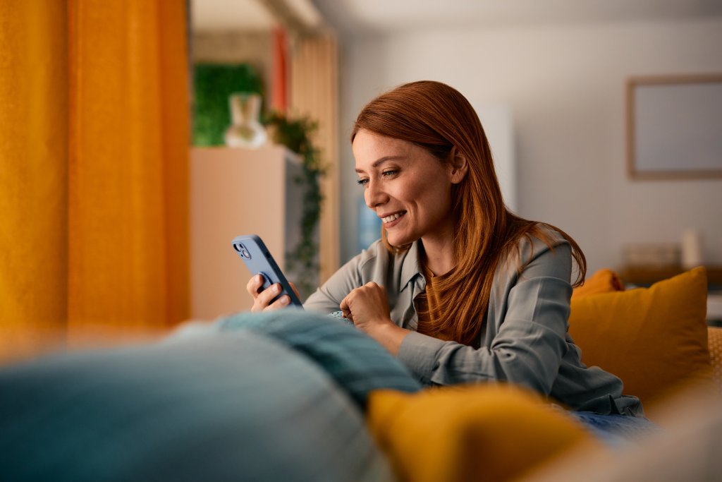 Frau nutzt Smartphone in gemütlicher Wohnzimmeratmosphäre, BLKB-Werbebild im Hintergrund