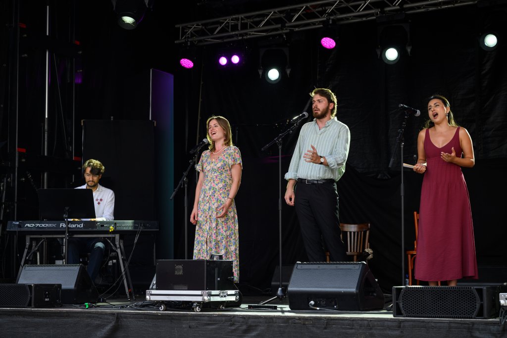 Vier Menschen stehen auf einer Bühne, einer spielt Keyboard, drei singen.