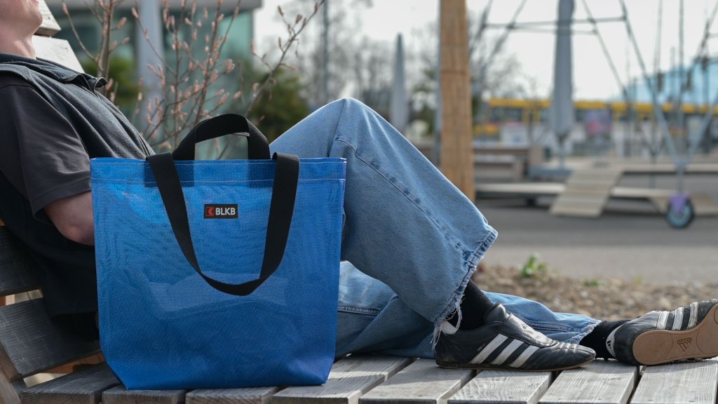 Eine Person sitzt auf einer Bank mit einer blauen Unikat-Tasche der BLKB an einem Park.