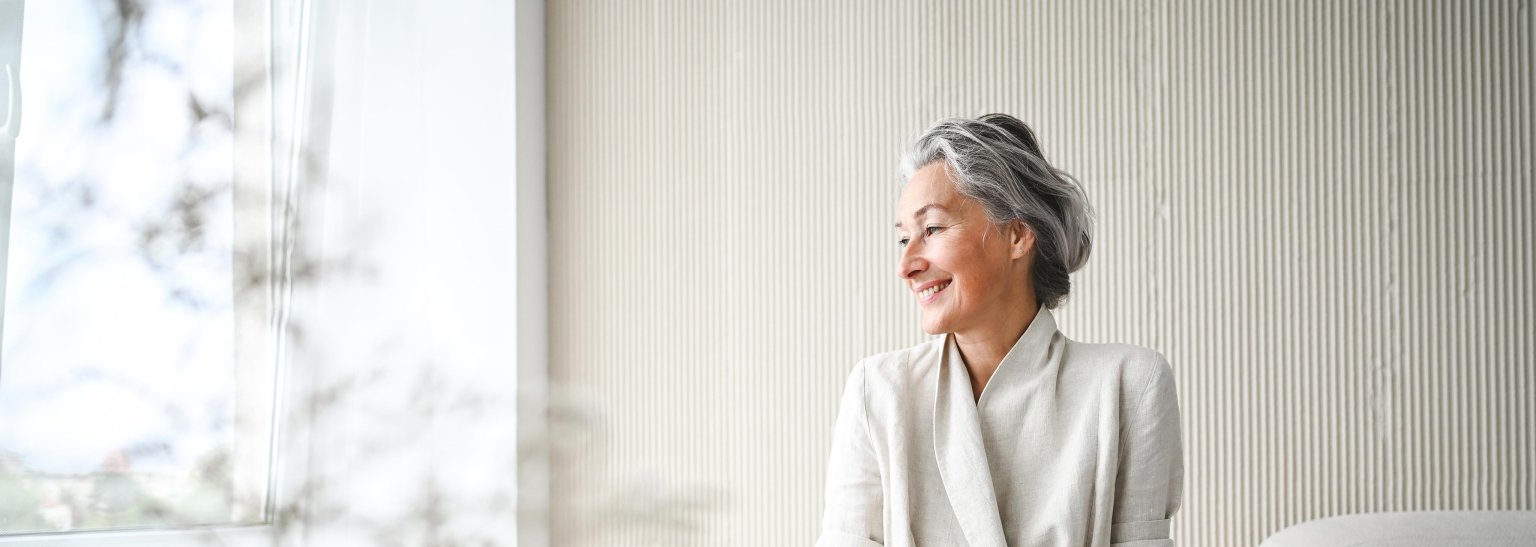 Ältere Frau sitzt entspannt im hellen Raum vor einem Fenster mit Sicht nach draussen