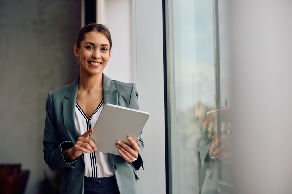 Eine Frau in einem Anzug hält ein Tablet und lächelt vor einem Fenster.